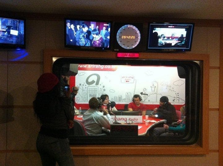 A live broadcast in progress at Arirang Radio's studio in Seoul, hosts at the red Arirang Radio desk under the ON AIR sign