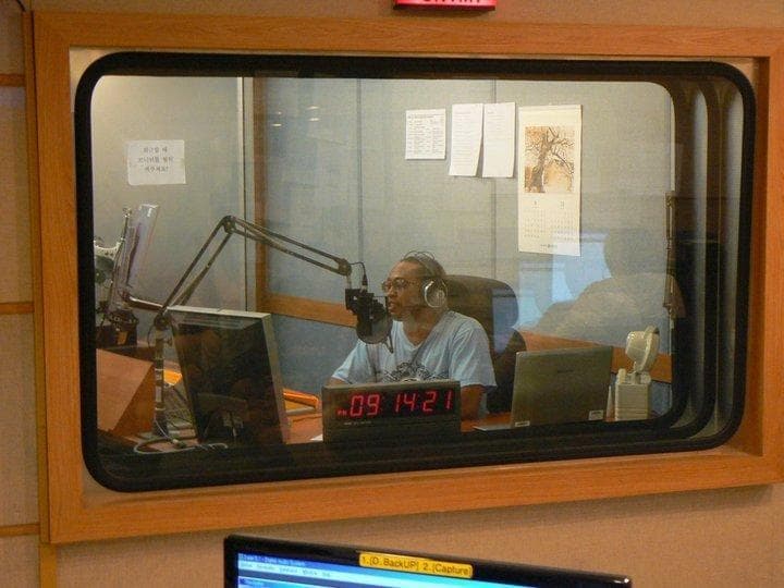 Roger Wong Won on the air behind the glass at Arirang Radio in Seoul, headphones on, at the mic, the on-air clock running
